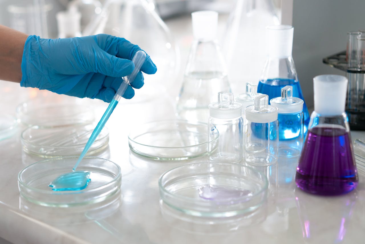 About Close-up of a scientists hand in glove conducting an experiment with petri dishes and colorful liquids in a lab.